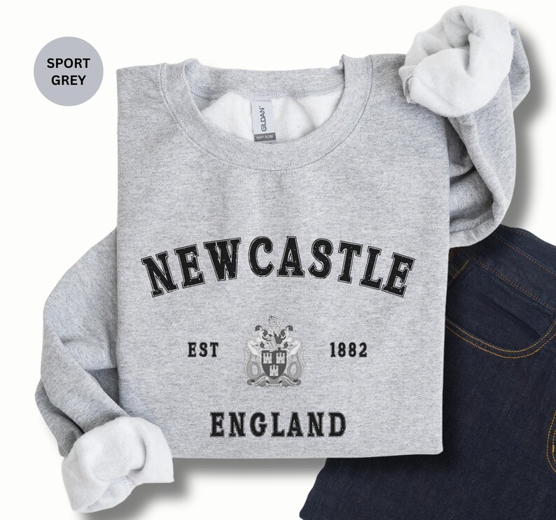 Lay flat sport grey sweatshirt with Newcastle  England, EST 1882 and the Newcastle coat of arms in White distressed text. Also in Black, Dark Heather, Forest Green, Navy, Sand, royal blue and White. Sizes from Small to XXL.