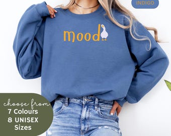 May include: A blue sweatshirt with the text "mood" in yellow and a white goose with an orange beak and feet. The sweatshirt is being worn by a person with long blonde hair.