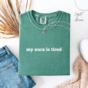 Flat lay Comfort Colors T Shirt in light green with ‘my aura is tired’ written in bold white lower case text