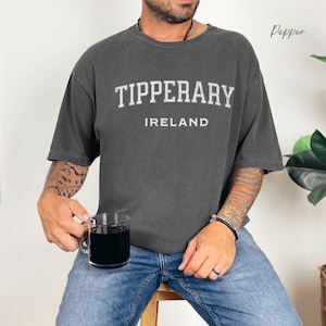 Man holding coffee cup wearing Pepper Comfort Colors® t-shirt  with Tipperary, Ireland in college style distressed look lettering.