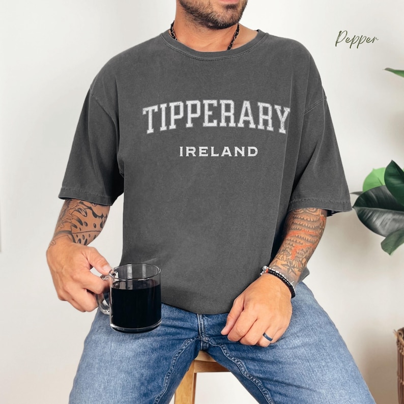 Man holding coffee cup wearing Pepper Comfort Colors® t-shirt  with Tipperary, Ireland in college style distressed look lettering.
