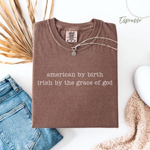 Flat lay of a folded espresso Comfort Colors T-shirt with the phrase “american by birth, irish by the grace of god” in typewriter-style text. Blue denim jeans on the left, brown dried leaves behind, a white trainer and cream blanket peeking in on the bottom left, with “espresso” labeled in the top right.