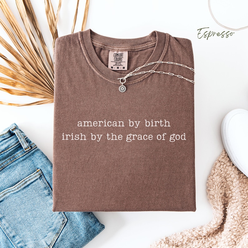 Flat lay of a folded espresso Comfort Colors T-shirt with the phrase “american by birth, irish by the grace of god” in typewriter-style text. Blue denim jeans on the left, brown dried leaves behind, a white trainer and cream blanket peeking in on the bottom left, with “espresso” labeled in the top right.