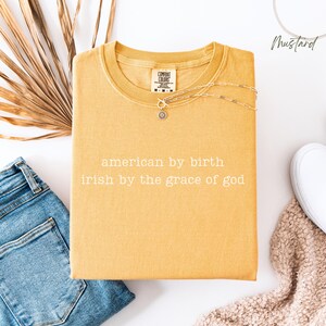 Flat lay showing a mustard Comfort Colors tee with “american by birth, irish by the grace of god” printed in a simple, lowercase font. Jeans to the left, dried foliage behind, plus cream blanket and white sneaker on the bottom. “Mustard” noted top right.