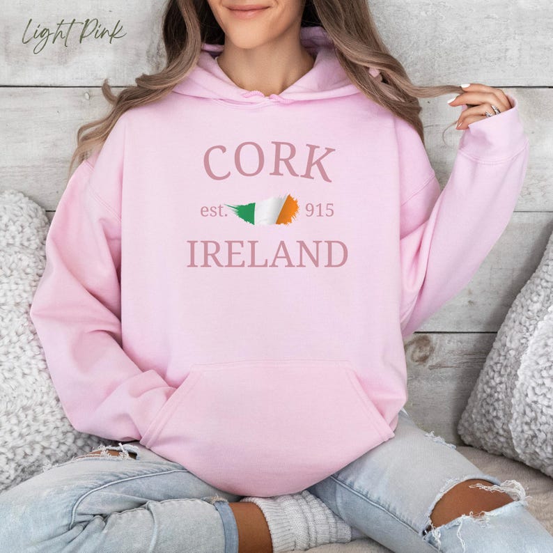 Woman wearing light pink hoodie Cork Ireland est 915 and Irish Flag. Sits casually cross legged, denim jeans, cozy socks against comfy cushions & a wooden light colored backdrop. 
Text is clean and bold. Aesthetic is relaxed and casual.