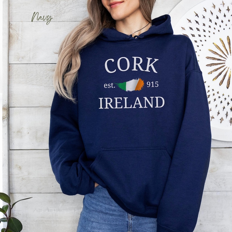 Woman wearing navy blue hoodie with Cork,  Ireland est. 915 in bold clean text stands casually  wearing denim jeans, against a light grey wooden backdrop. Customize with your hometown.  Neutral casual vibe.