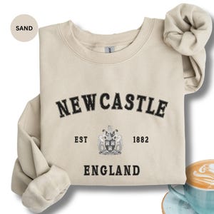 Lay flat sand sweatshirt with Newcastle  England, EST 1882 and the Newcastle coat of arms in White distressed text. Also in Black, Dark Heather, Forest Green, Navy, royal blue, Sport Grey and White. Sizes from Small to XXL.