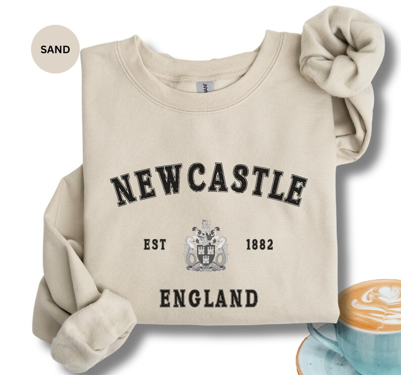 Lay flat sand sweatshirt with Newcastle  England, EST 1882 and the Newcastle coat of arms in White distressed text. Also in Black, Dark Heather, Forest Green, Navy, royal blue, Sport Grey and White. Sizes from Small to XXL.
