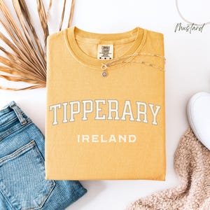 Flat Lay folded comfort colors® T shirt in Mustard with Tipperary, Ireland in college style distressed look lettering. The shirt is in the centre of the picture with a pair of blue jeans to the left, some brown dried foliage sitting to the back left of the image.