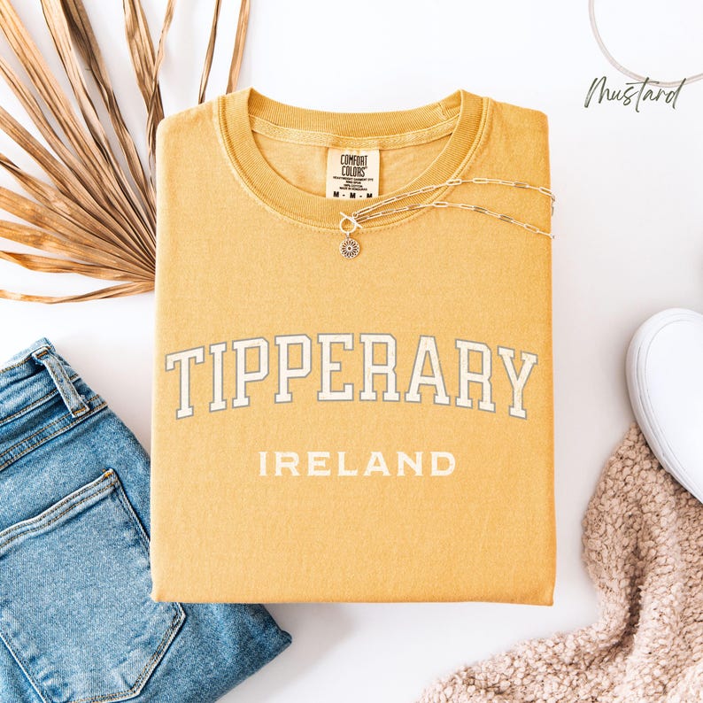 Flat Lay folded comfort colors® T shirt in Mustard with Tipperary, Ireland in college style distressed look lettering. The shirt is in the centre of the picture with a pair of blue jeans to the left, some brown dried foliage sitting to the back left of the image.