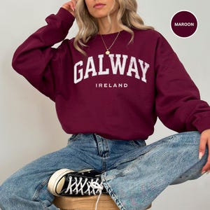 Woman wears maroon burgundy sweatshirt with Galway, Ireland in a forest green vintage style text. Option to customise.