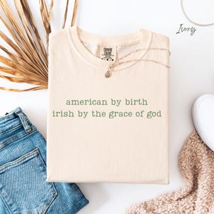 Folded natural Comfort Colors T-shirt with lowercase typewriter quote “american by birth, irish by the grace of god.” Styled with denim jeans, dried leaves, a soft cream blanket and a white trainer. “Natural” color label shown at the top right.