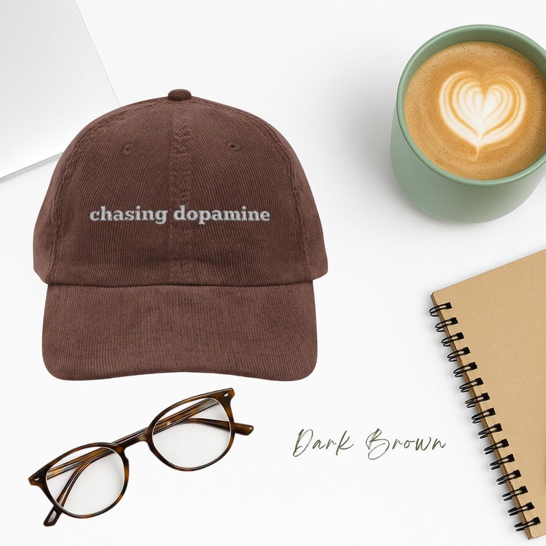 Vintage Corduroy dad hat in dark brown with embroidered text “chasing dopamine” in light grey thread . A minimalist, mental health inspired design set against a desktop scene of coffee , laptop, reading glasses & notebook Available in multiple colors.