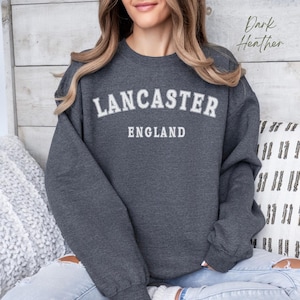 Pode incluir: Uma camisa de moletom cinza escuro com o texto "LANCASTER ENGLAND" em branco.