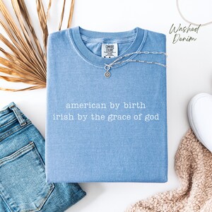 Washed denim Comfort Colors shirt folded neatly, displaying “american by birth, irish by the grace of god” in small typewriter text. Paired with jeans, dried leaf accents, white shoe, and cream blanket. Color name “washed denim” noted at top right.