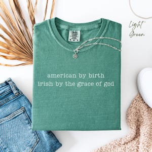 Folded light green Comfort Colors T-shirt with “american by birth, irish by the grace of god” in typewriter font. Surrounded by denim on the left, dry leaves behind, a sneaker and soft cream blanket in the corner. “Light green” text appears top right.