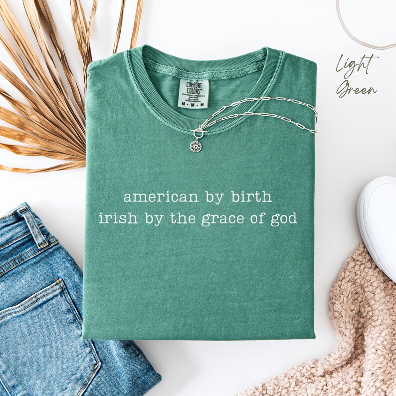Folded light green Comfort Colors T-shirt with “american by birth, irish by the grace of god” in typewriter font. Surrounded by denim on the left, dry leaves behind, a sneaker and soft cream blanket in the corner. “Light green” text appears top right.