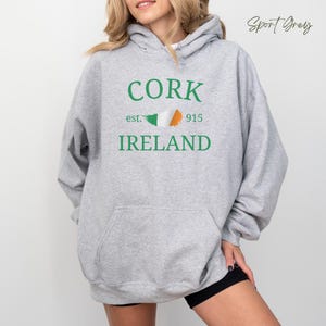 Woman wearing sport grey hoodie with Cork,  Ireland est. 915 in bold clean text stands casually  wearing shorts, against a light grey wooden backdrop. Customize with your hometown.  Neutral casual vibe.