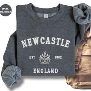 Lay flat dark heather sweatshirt with Newcastle  England, EST 1882 and the Newcastle coat of arms in White distressed text. Also in Black, Royal,  Forest Green, Navy, Sand, Sport Grey and White. Sizes from Small to XXL.