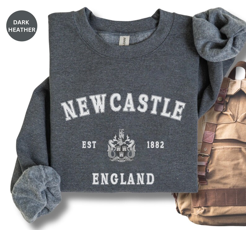 Lay flat dark heather sweatshirt with Newcastle  England, EST 1882 and the Newcastle coat of arms in White distressed text. Also in Black, Royal,  Forest Green, Navy, Sand, Sport Grey and White. Sizes from Small to XXL.