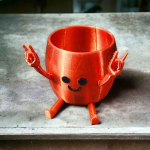May include: A 3D printed orange planter shaped like a cartoon character with arms and legs. The character has a smiling face with two black eyes and is making the rock and roll hand gesture with both hands.