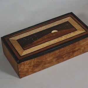May include: A wooden box with a multi-layered inlay design on the lid. The inlay depicts a night sky with stars, a crescent moon, and a mountain range. The box is made of various wood types, including dark wood, light wood, and a reddish-brown wood.
