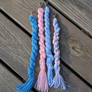 Op de afbeelding: Vier handgemaakte sleutelhangers met gevlochten garen in blauwe en roze tinten. Elke sleutelhanger heeft een zilverkleurige sluiting en een kwast van los garen aan de onderkant. De sleutelhangers zijn gerangschikt op een verweerd houten oppervlak.