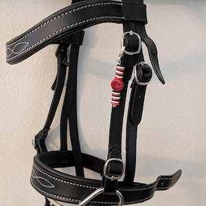 Puede incluir: Un cabezal de caballo de cuero negro con costuras blancas y herrajes plateados. Un charm decorativo con cuentas rojas, blancas y negras y una carita sonriente está adjunto. Diseñado para uso ecuestre.