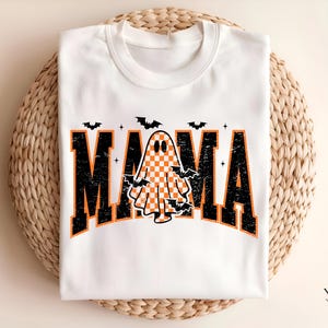 May include: White crew neck t-shirt with a Halloween-themed design. The word "MAMA" is printed in black with orange accents, featuring a checkered ghost and bats. The shirt is displayed on a woven surface.