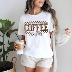 May include: A white t-shirt with the words "COFFEE weather" in brown, checkered font. The person is holding a glass of iced coffee. The shirt is paired with beige shorts. A beige cardigan is draped over the person's shoulders.