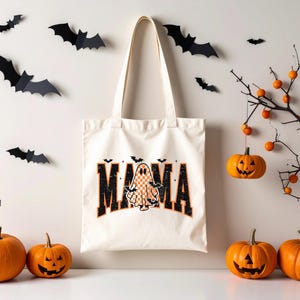 May include: A cream-colored tote bag with the word "MAMA" in black and orange lettering, featuring a ghost and bat graphic. The Halloween-themed image includes orange pumpkins, paper bats, and a decorative branch with hanging pumpkins.