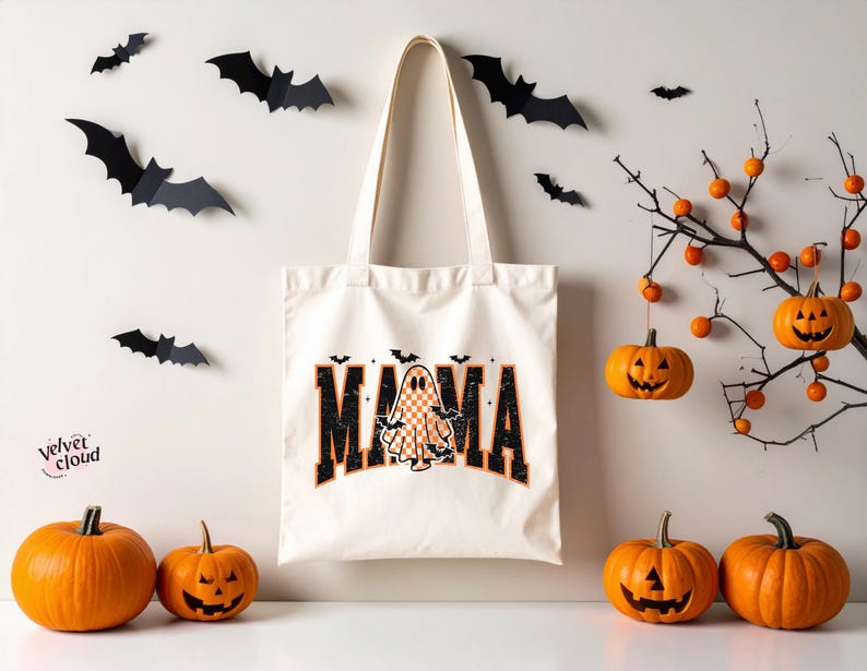 May include: A cream-colored tote bag with the word "MAMA" in black and orange lettering, featuring a ghost and bat graphic. The Halloween-themed image includes orange pumpkins, paper bats, and a decorative branch with hanging pumpkins.