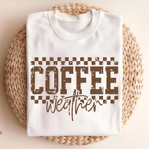 May include: A white t-shirt with the words "COFFEE weather" in brown, distressed font. The text is framed by a checkered pattern. The shirt is displayed on a woven, circular placemat.