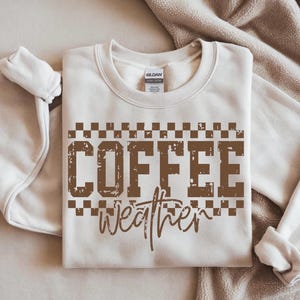 May include: Cream-colored sweatshirt with the words "COFFEE weather" in brown, distressed font. The word "COFFEE" is in a block letter style, and the word "weather" is in a cursive font. The sweatshirt has a crew neck and long sleeves.