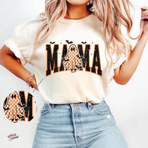 May include: Cream-colored t-shirt with the word "MAMA" in black block letters, featuring a checkered ghost graphic. Bats are above and around the word. The shirt is a Halloween-themed design.
