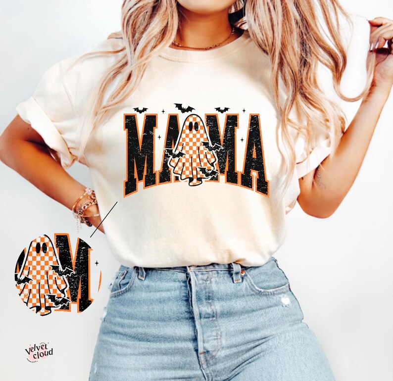 May include: Cream-colored t-shirt with the word "MAMA" in black block letters, featuring a checkered ghost graphic. Bats are above and around the word. The shirt is a Halloween-themed design.