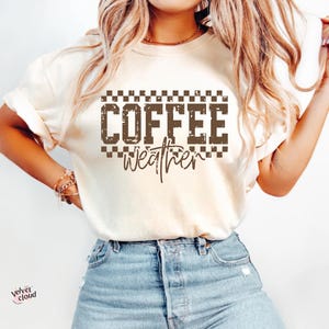 May include: Cream-colored t-shirt with the words "COFFEE weather" in a brown, distressed font. The text is framed by a checkered pattern. The shirt is paired with blue denim jeans.