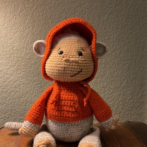 May include: A handmade crochet monkey doll with a light brown face and ears, wearing an orange hoodie. The monkey has a gray body and limbs, and is sitting on a wooden surface. The doll is a soft toy.
