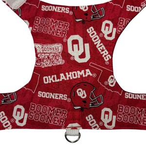 May include: Red dog harness featuring the words "SOONERS", "BOOMER SOONER", and "OKLAHOMA" in white. The harness has a repeating pattern of football helmets and the Oklahoma Sooners logo. A metal D-ring is attached.