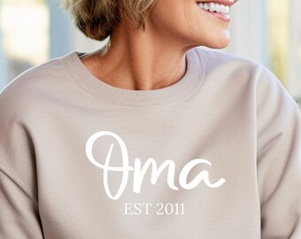 May include: A beige long-sleeve crewneck sweatshirt featuring the white text "Oma" in a large script font, with the smaller white text "EST 2011" printed directly underneath the main word. The garment has a simple, solid-colored design.