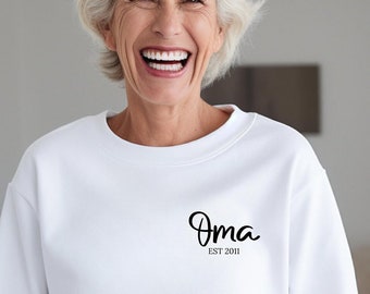 Puede incluir: Una sudadera blanca de manga larga con cuello redondo que presenta el texto negro "Oma" en una fuente cursiva en la parte superior izquierda del pecho, con el texto "EST 2011" impreso en una fuente sans-serif más pequeña debajo de "Oma."