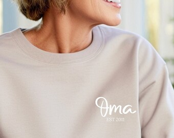 May include: A beige crewneck sweatshirt featuring the white text "Oma" in a cursive script font on the upper left chest, with the smaller white text "EST 2011" printed directly underneath the word "Oma" in a clean, sans-serif typeface.