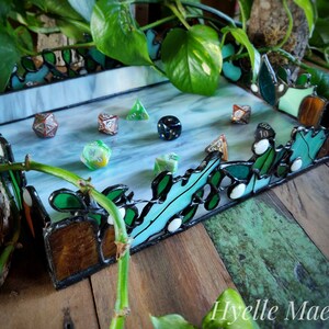 May include: A stained glass dice tray with a light blue interior and a border of green and brown glass. Several polyhedral dice are scattered across the tray. The tray is decorated with green leaf-like designs. A candle sits in a wooden holder.