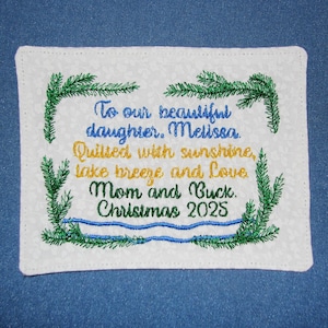 May include: A rectangular white fabric patch with embroidered green pine branches framing the text: To our beautiful daughter, Melissa. Quilted with sunshine, lake breeze and Love. Mom and Buck. Christmas 2025. The text is in blue, gold, and green.