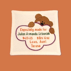 May include: Embroidered square baby keepsake with a brown teddy bear peeking over a purple-outlined cloud shape. The cloud contains the text "Especially made for Julian Armando Urbaniak, 9-25-25, 8 lbs 12 oz, Love, Aunt Teresa."