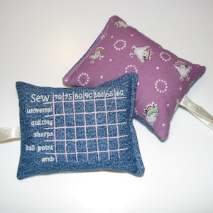 May include: Two small fabric pincushions. One is blue denim with a white grid and the text "Sew 70 75 80 90 100 105 160 universal quilting sharps ball point emb". The other is purple with white polka dots and a white bird design.