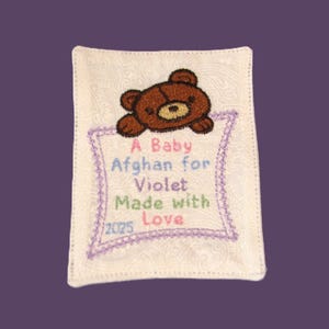 May include: Embroidered baby afghan with a brown teddy bear and the text "A Baby Afghan for Violet Made with Love 2025" in colorful thread. The afghan is white with a paisley pattern and a purple border.