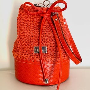 Könnte beinhalten: Orangener Eimerbeutel mit gewebtem Oberteil und strukturierter Unterseite. Die Tasche hat einen roten Lederkordelverschluss und einen passenden Schulterriemen. Die Tasche hat silberne Hardware und den Text "Handmade. Timeless. Unstoppable."