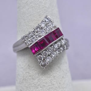 May include: A silver ring featuring a geometric design with a central row of three square-cut, vibrant red gemstones. The ring is flanked by rows of small, sparkling white gemstones, creating a striking contrast. The ring is set on a light-colored background.