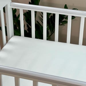 May include: A white cot with a white mattress. The cot has vertical bars and a rounded top rail. Green leafy plants are visible behind the cot.